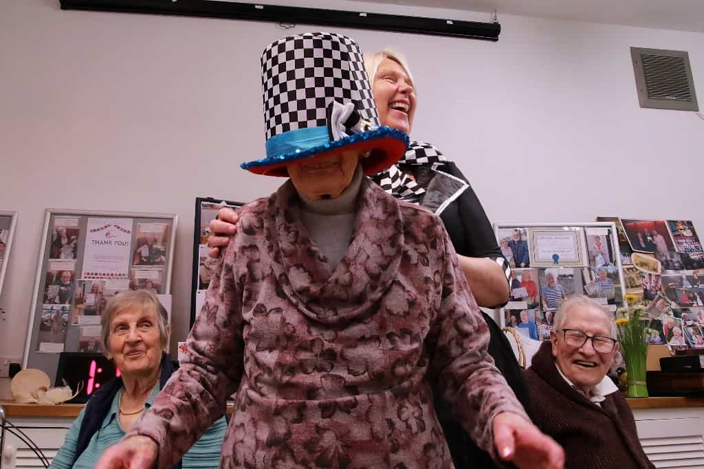 Bebe wears an oversized Mad Hatter's hat that has slipped over her face, while manager Fiona laughs behind her.