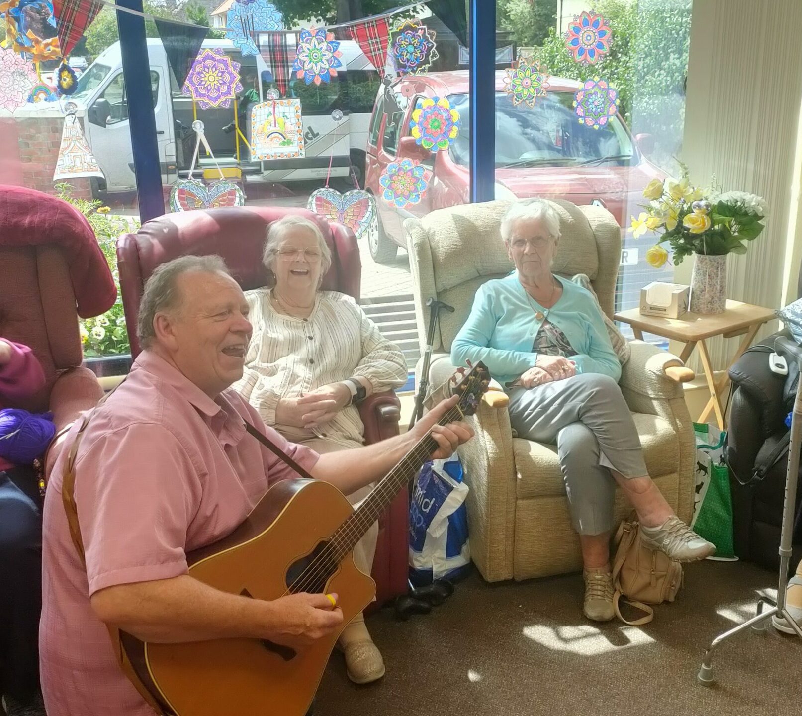 Alex Hodgson plays acoustic guitar and sings for two seated Harlawhill members by the window. One laughs while the other enjoys the music.